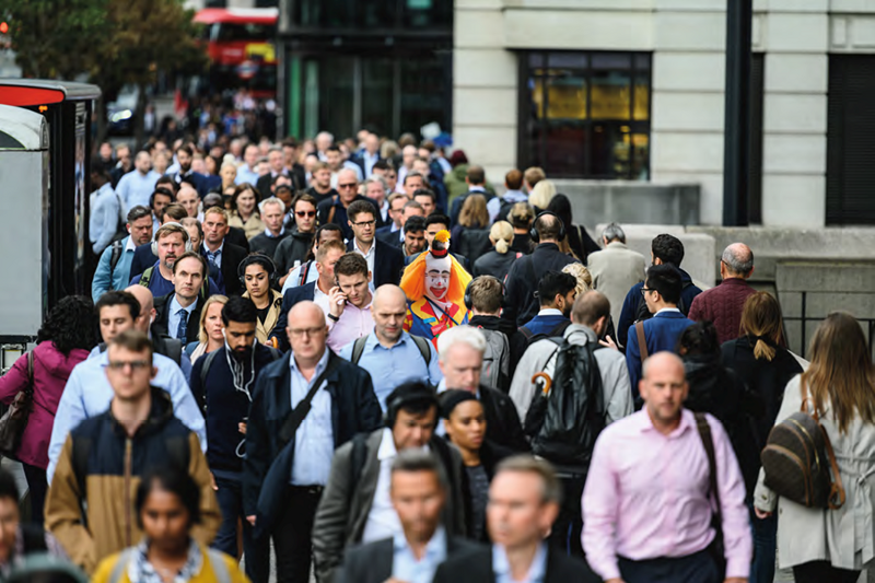A crowd of people walking along a pavement, with a clown amongst the group